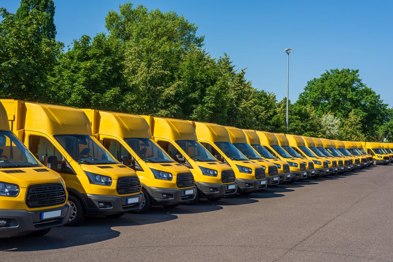 A fleet of electric yellow delivery vans are parking in front of green trees.